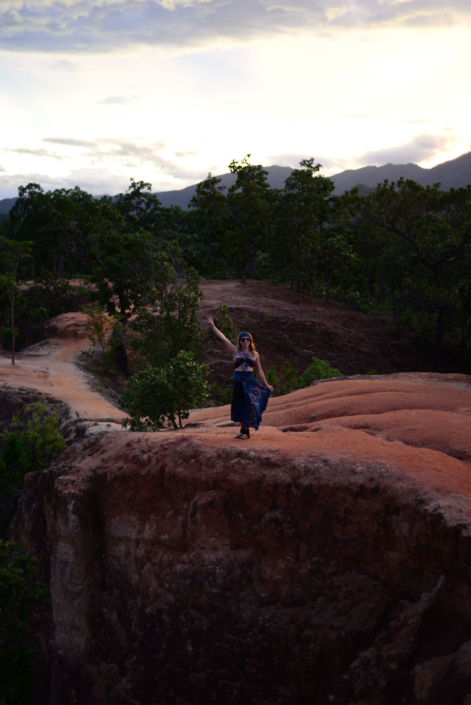 Silhouetted figure stands atop red rock formation at sunset with mountains and trees in background landscape.