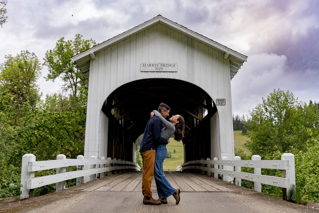 Couple sharing a romantic kiss in front of a white covered bridge surrounded by lush green trees.