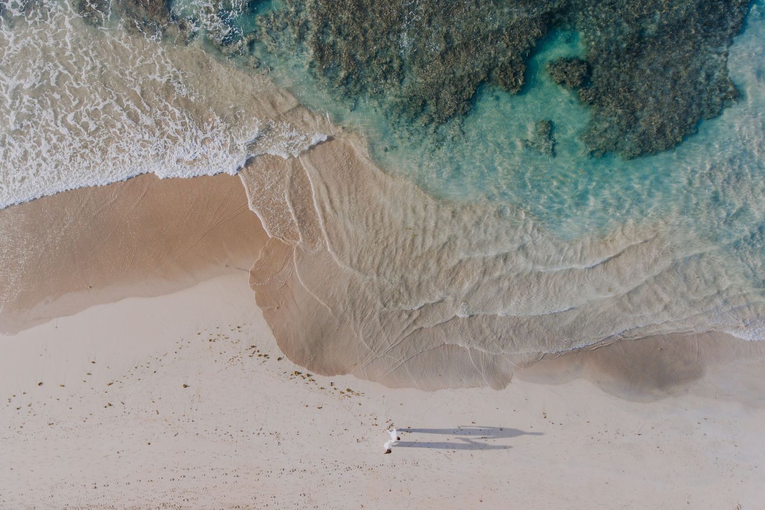 Aerial view of turquoise ocean waves meeting white sandy beach with small shadows visible.