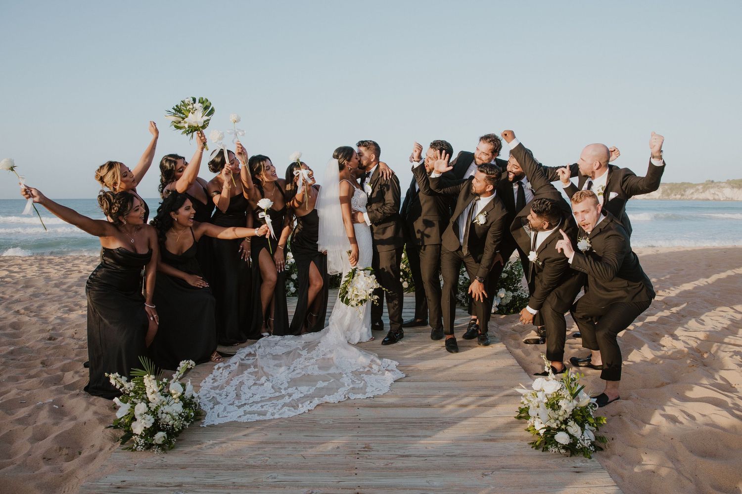 Bridal party celebrates on beach with joyful poses at sunset.