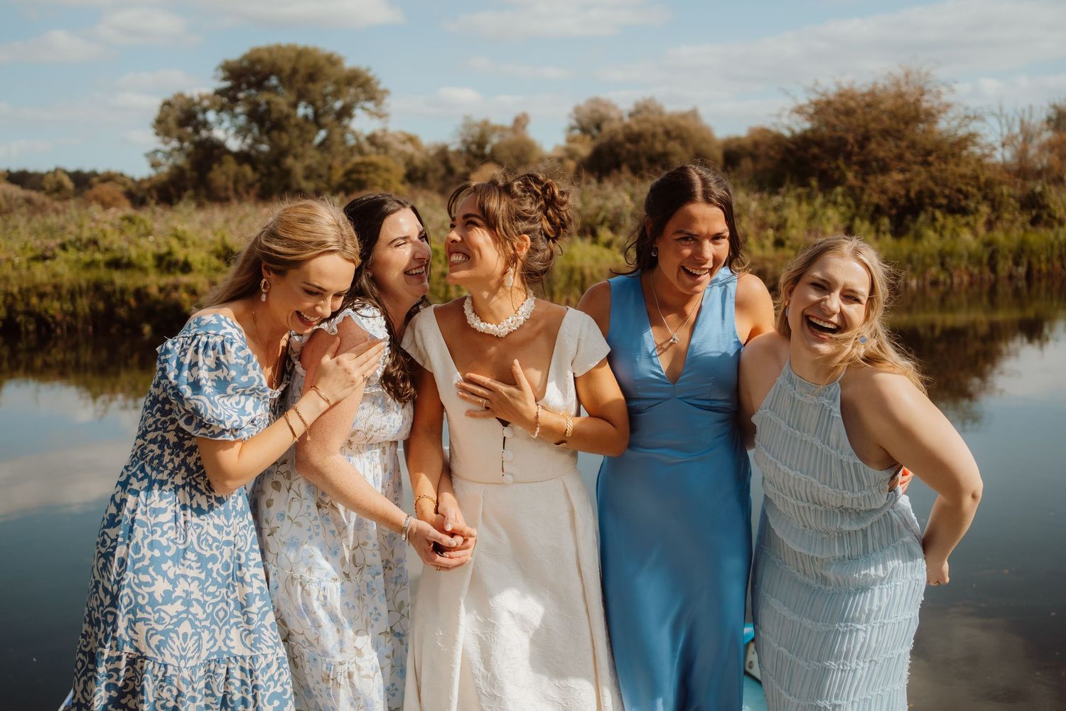 A group of friends in summer dresses laughing together by a serene lake during sunset.