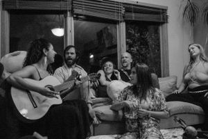 A group gathers in a living room for an acoustic guitar singalong session in black and white.
