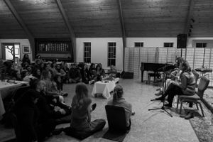 An audience sits on the floor listening to performers in a large room with wooden beams.