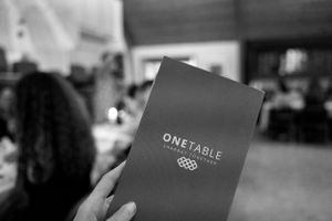 A hand holds a OneTable branded menu or program against a blurred background.
