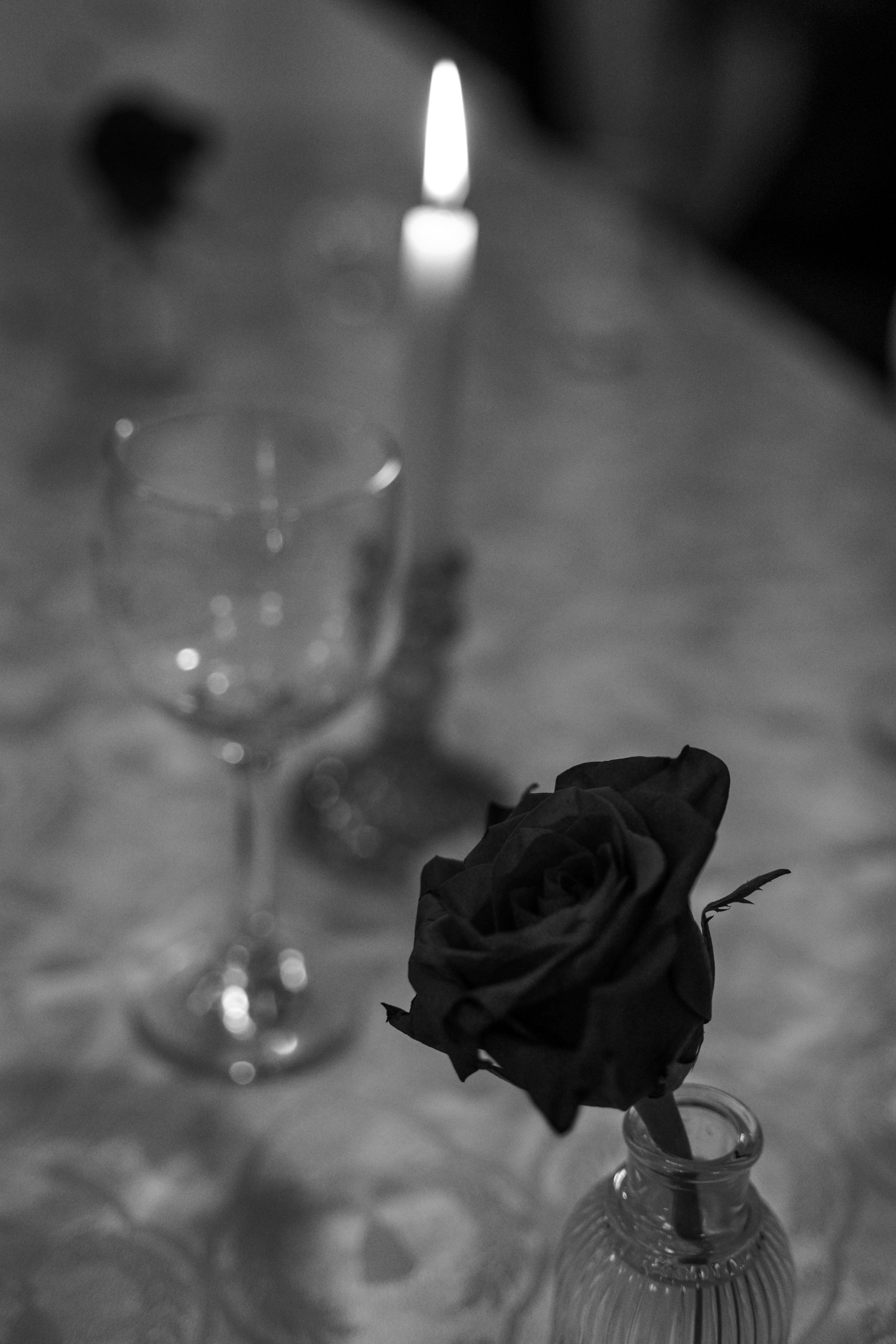 Moody black and white still life with a lit candle, wine glass, and dark rose in a vase.