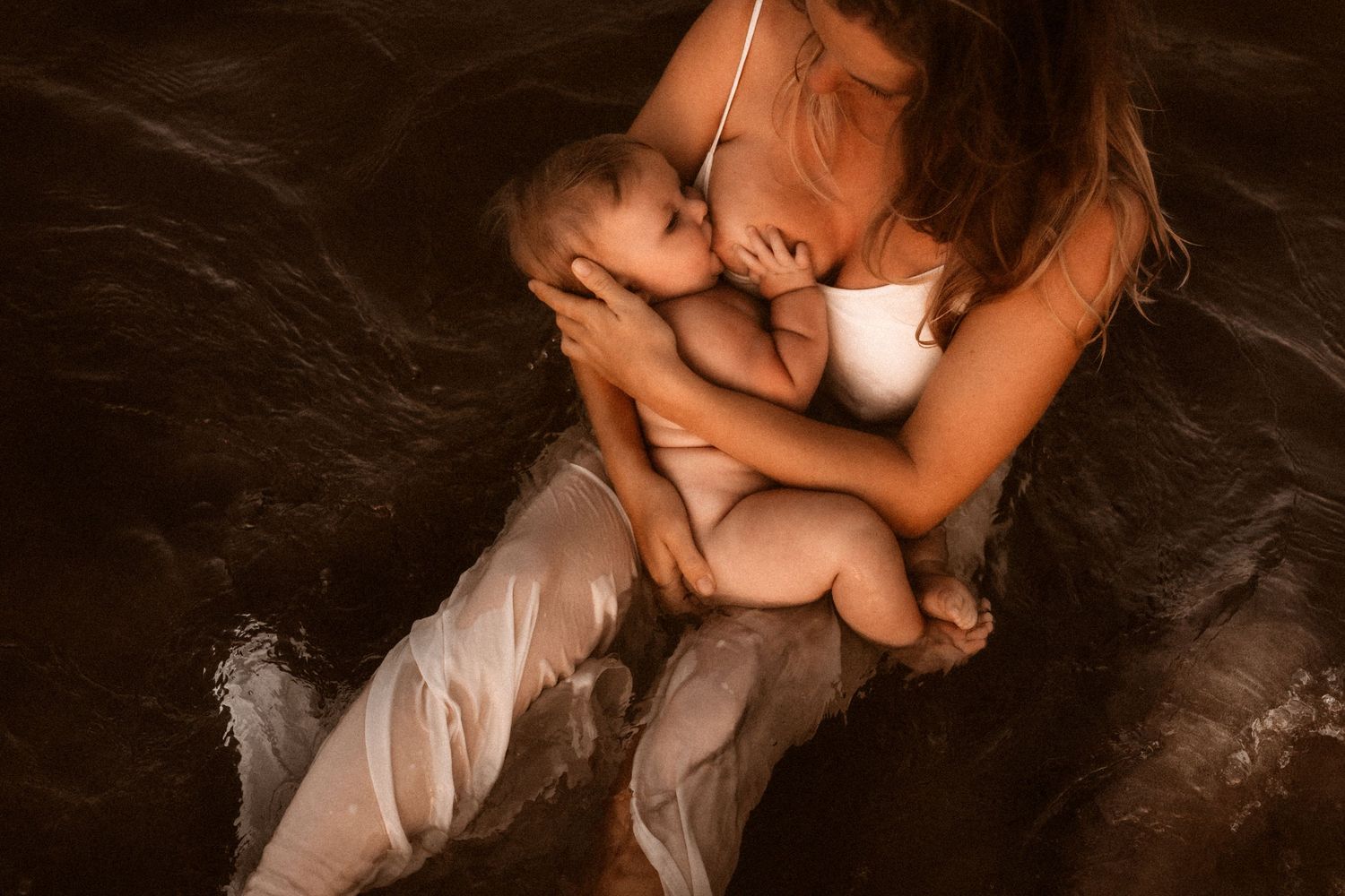 An intimate photo showing tender skin-to-skin contact between parent and baby in dark artistic lighting. Photographer Javea