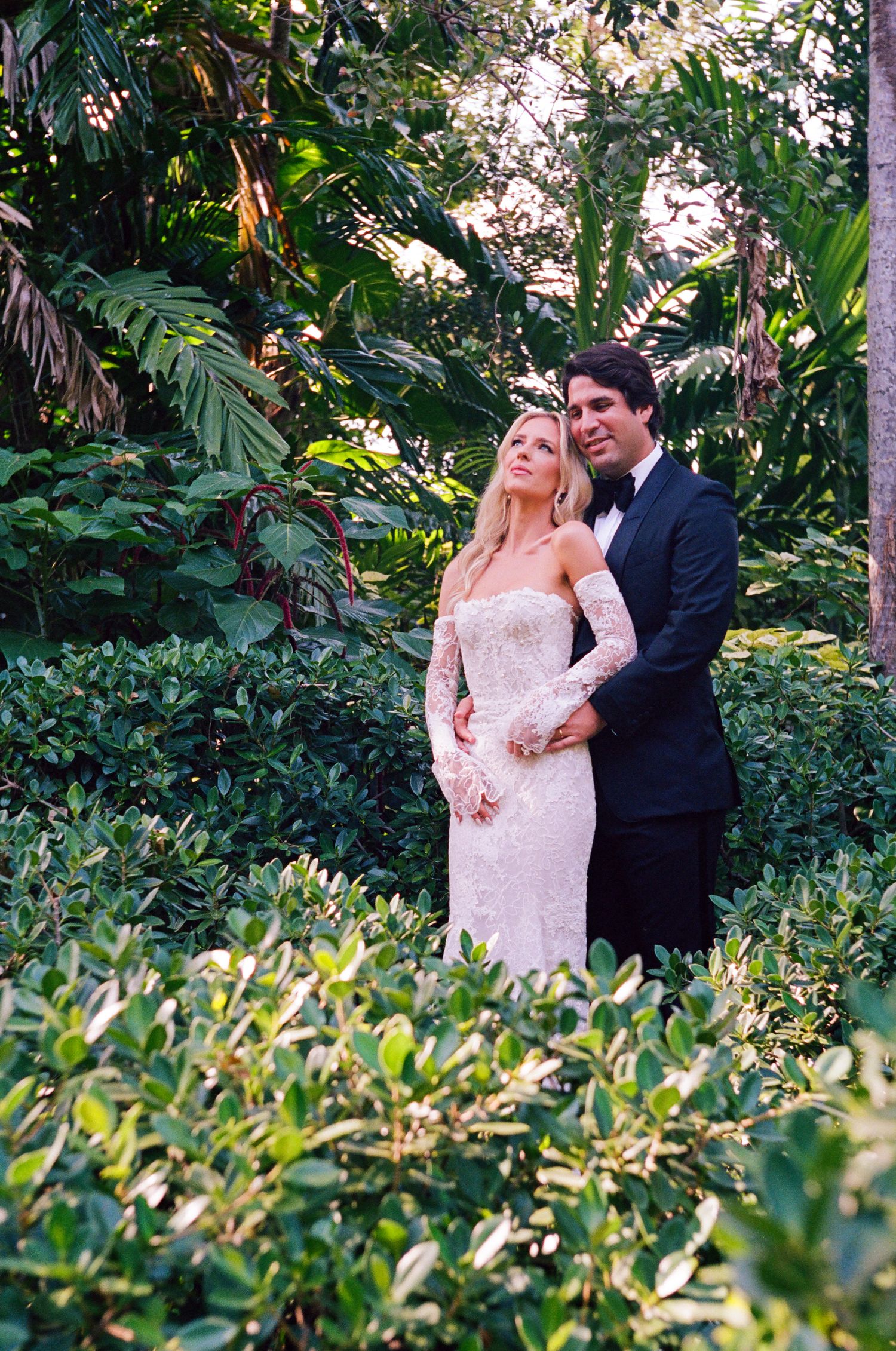 A couple in wedding attire pose lovingly together among lush green foliage in a garden setting.