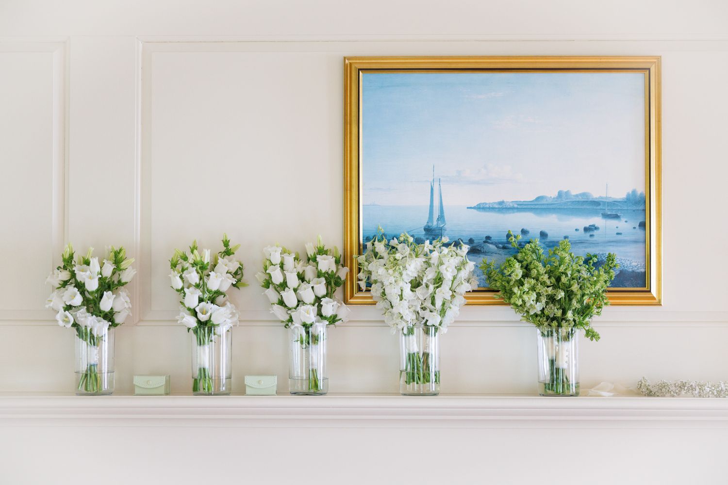 White floral arrangements in glass vases displayed on a mantel with framed coastal artwork.