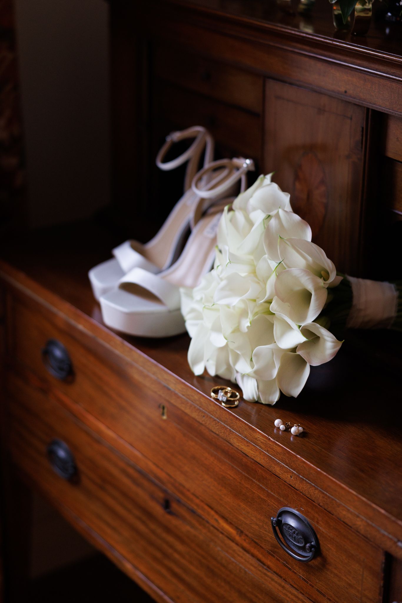 White bridal bouquet and wedding shoes arranged on a vintage wooden dresser.