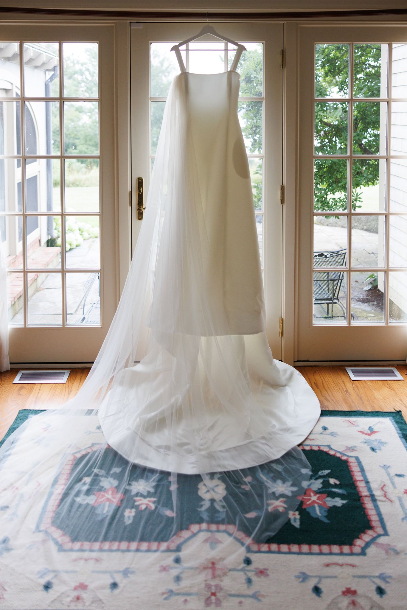 A flowing white wedding dress displayed on a patterned blue and pink rug near large windows.