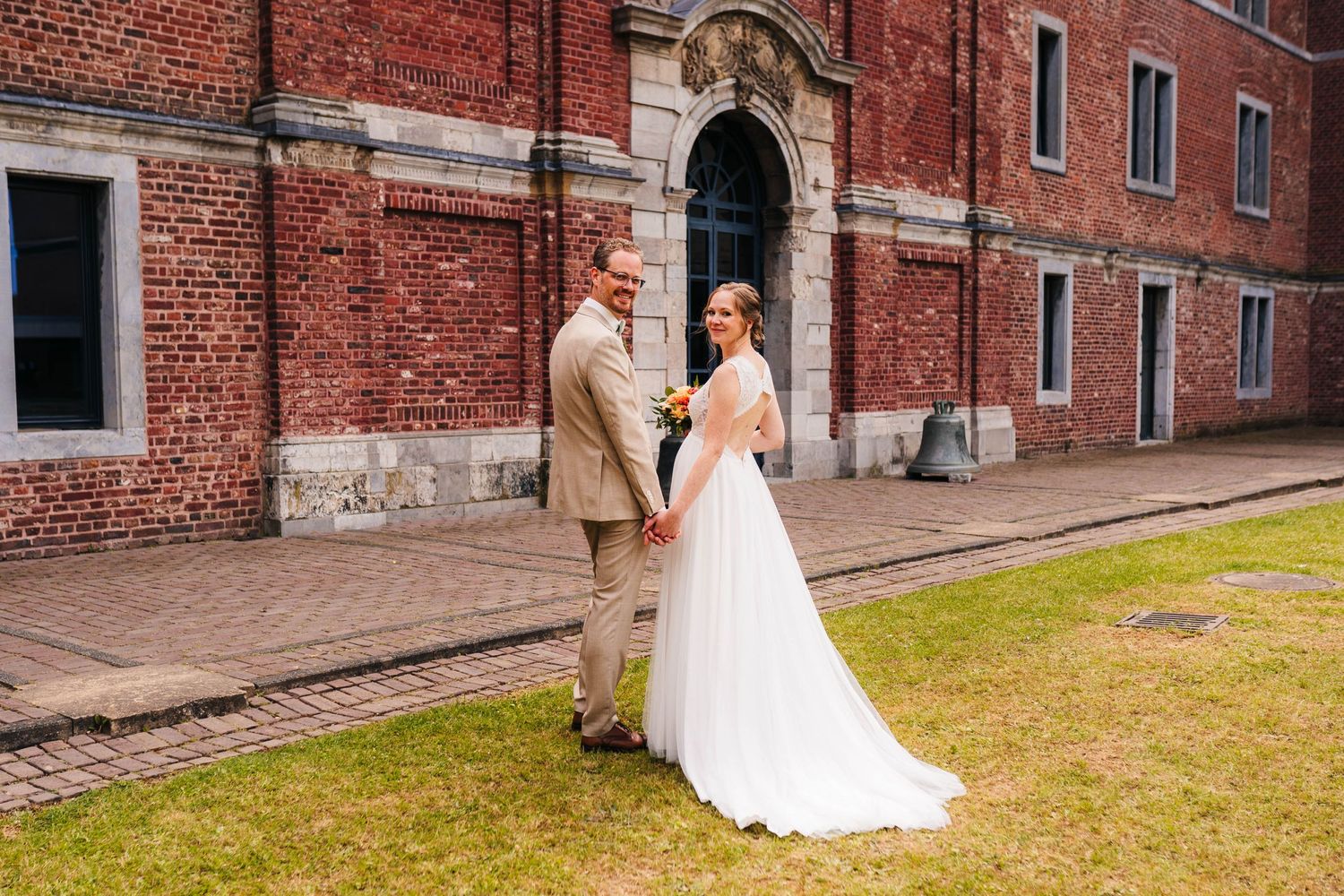 A couple stands together on their wedding day in front of a historic red brick building.