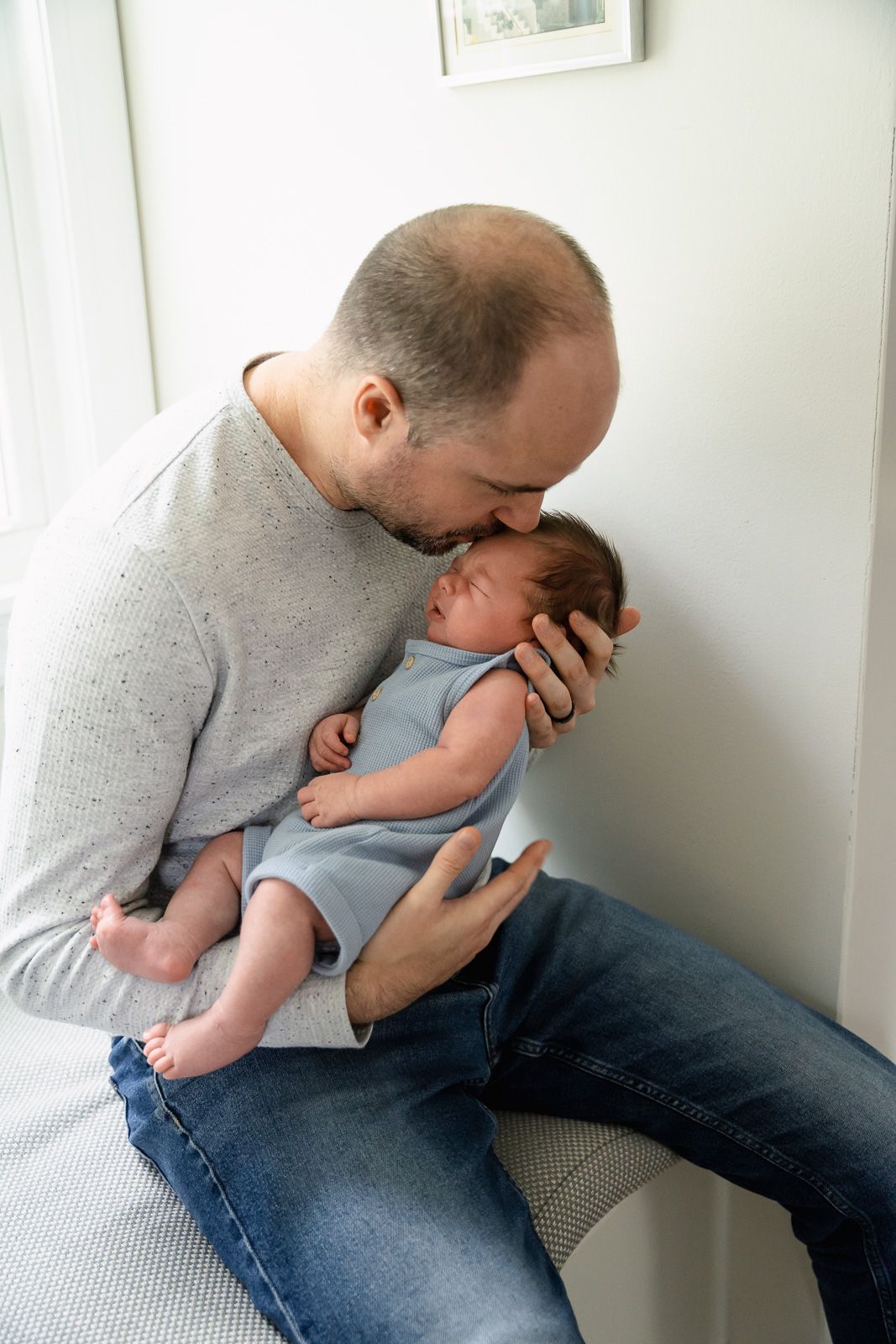 A tender moment shows a parent in gray sweater hugging and kissing a baby wearing blue romper by a sunny window in Arlington Heights