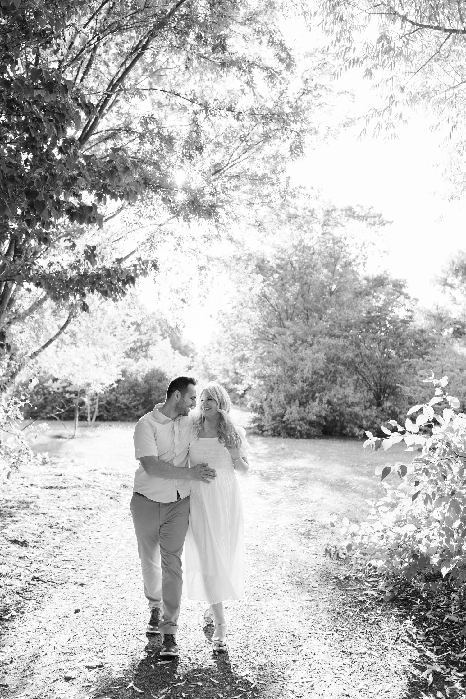 A couple in light-colored clothing walks together on a sunlit path through trees and foliage in a dreamy black and white photo.