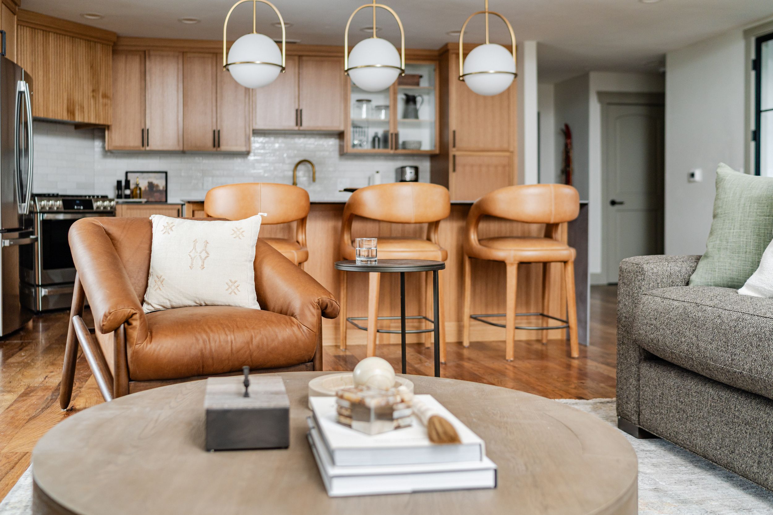 Modern open concept living space with leather armchair, round coffee table, and wooden kitchen featuring pendant lighting.