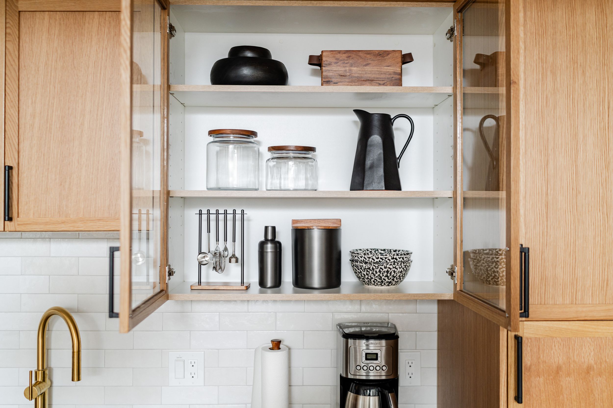 Modern kitchen cabinet with wooden doors displaying organized coffee station, glass jars, and black appliances.