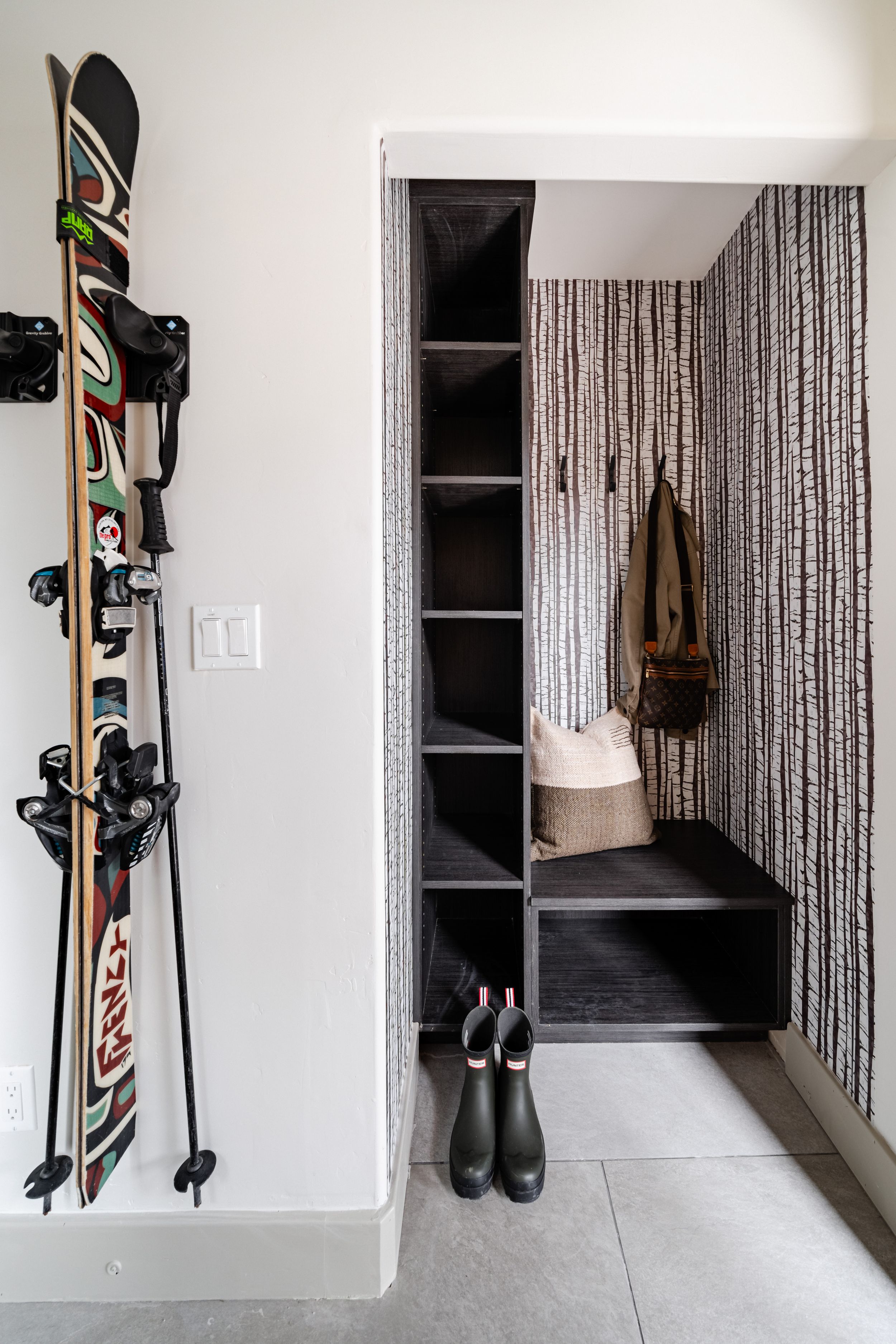 Modern entryway with mounted snowboard, built-in shelving unit, and black boots on gray tile flooring.