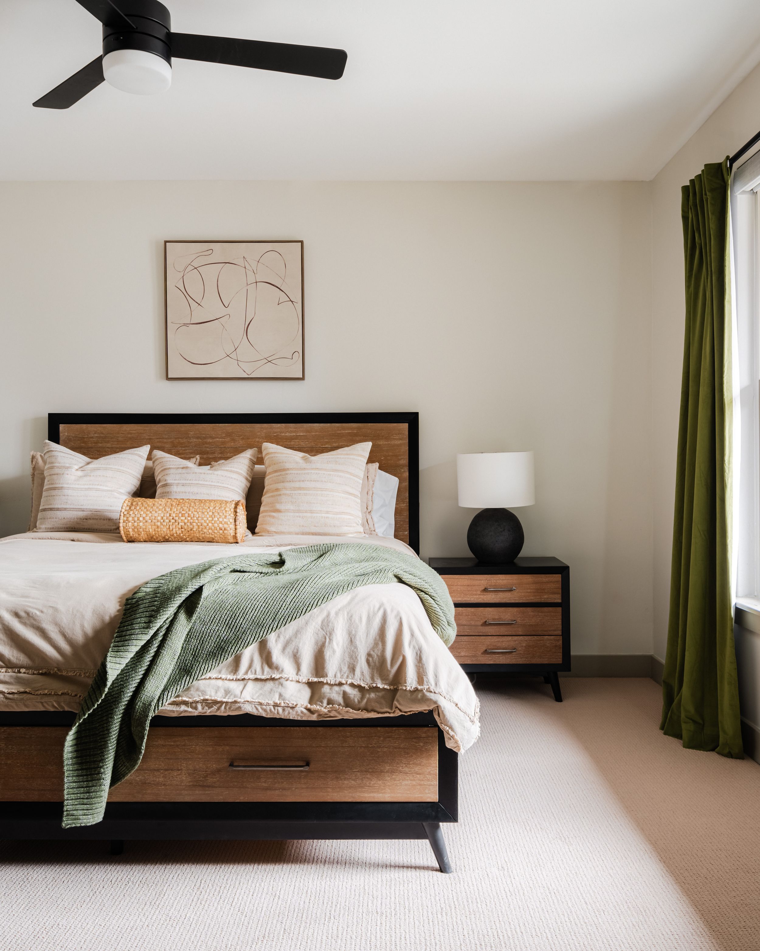 Modern bedroom with wooden bed frame, green curtains, abstract wall art, and round black table lamp.