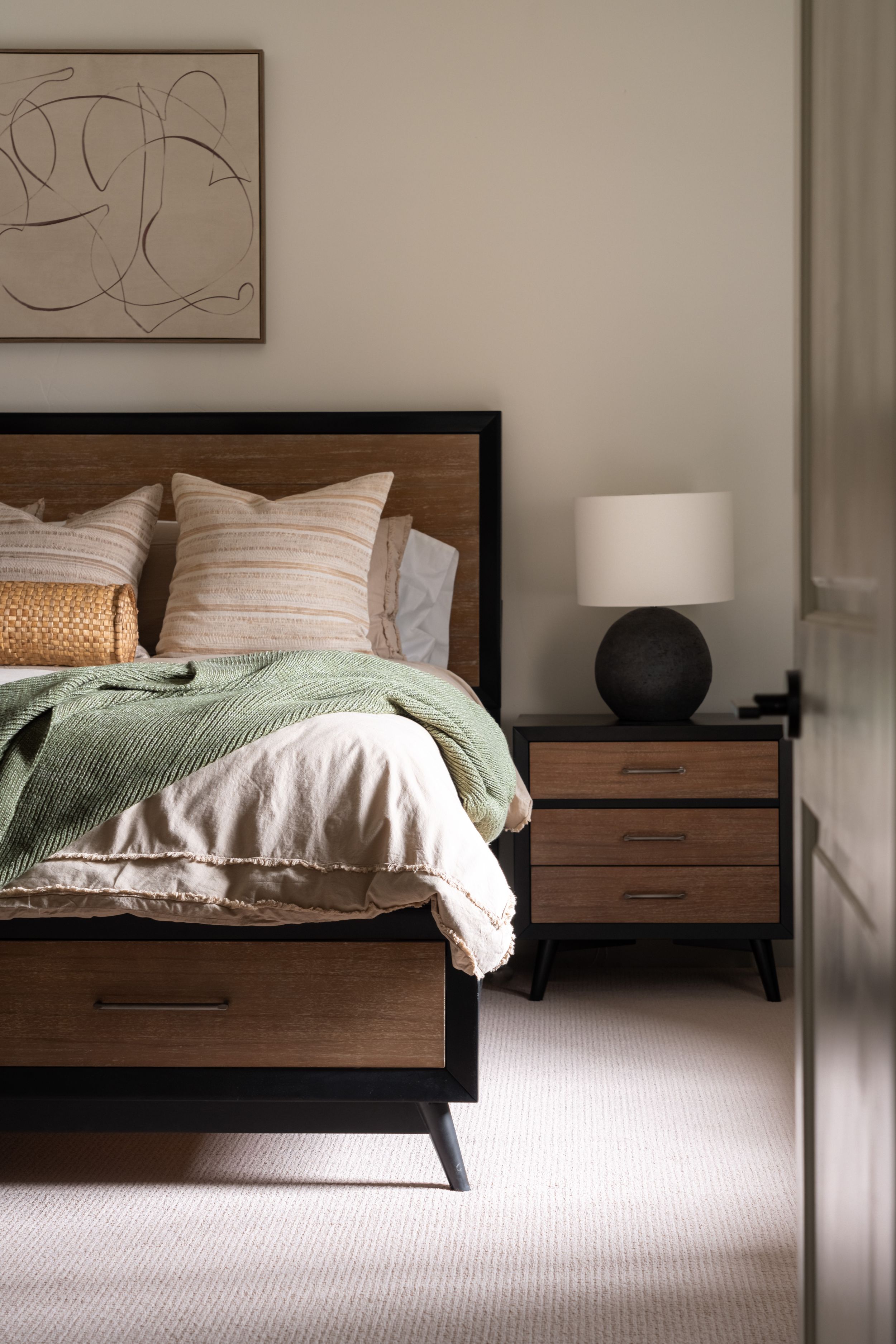 Modern bedroom featuring a wooden bed frame with white bedding, sage green throw, and round black table lamp on nightstand.