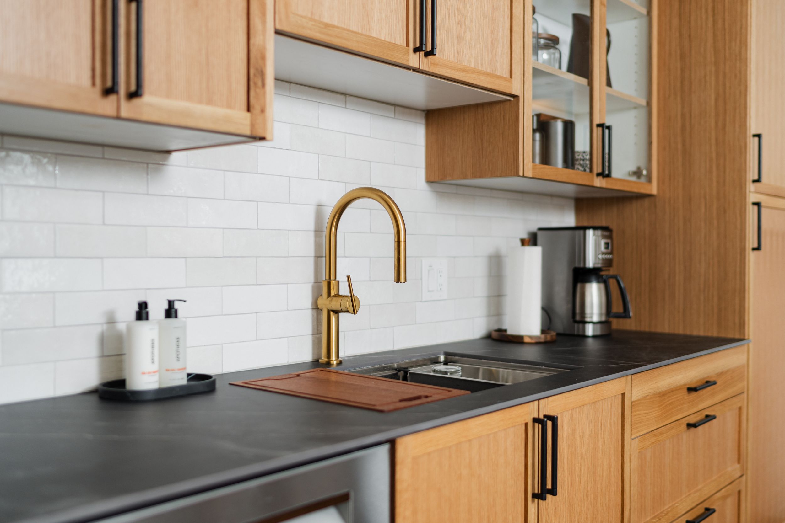 Modern kitchen with light wood cabinets, brass hardware, white subway tile backsplash, and dark countertops.