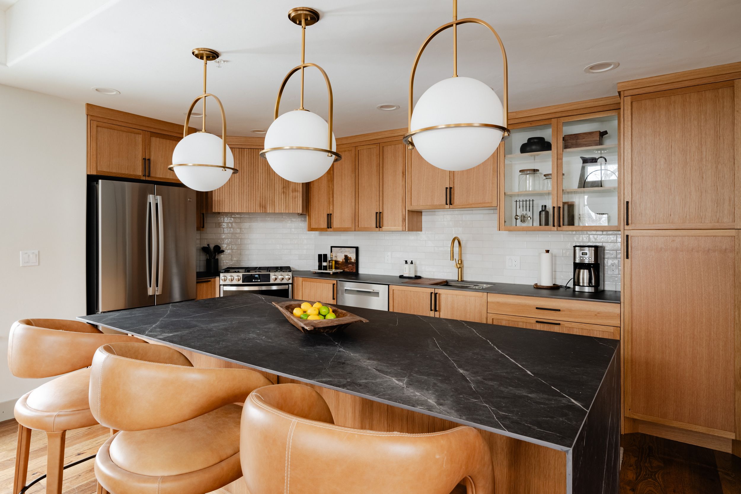 Modern kitchen featuring light wood cabinets, marble black countertops, globe pendant lights, and leather bar stools.