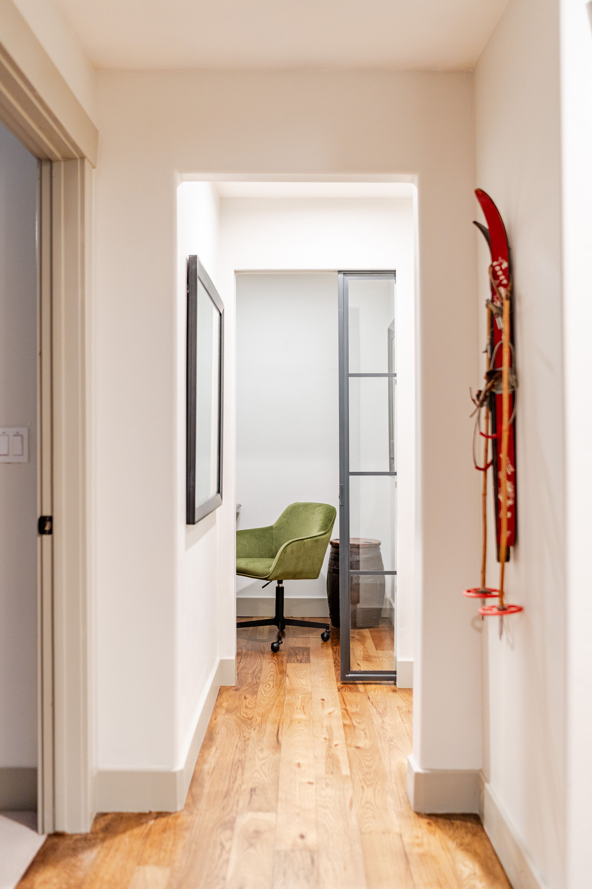 Minimalist hallway with mounted red ski on white wall leading to home office with wood flooring and green chair.