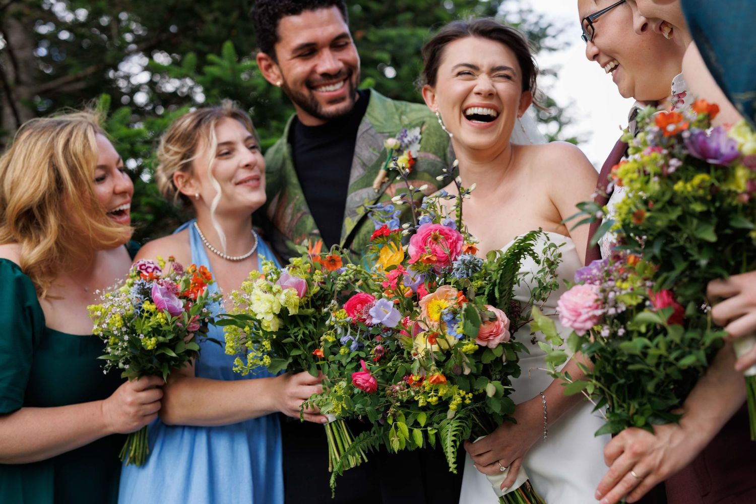 Wedding party holding vibrant wildflower bouquets while laughing together outdoors.