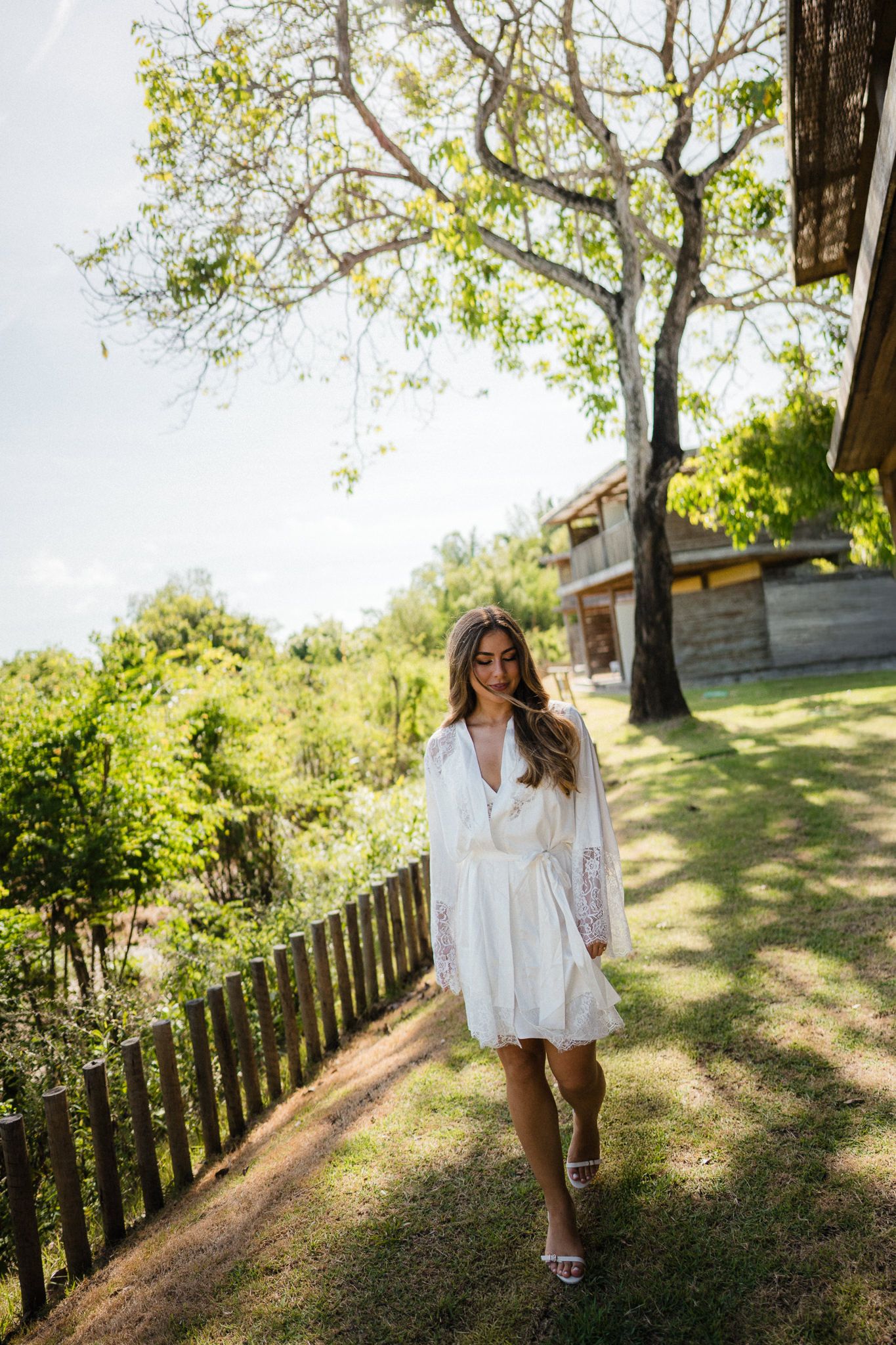 Person in flowy white dress walks gracefully through a sunlit garden path with trees and a fence in the background.