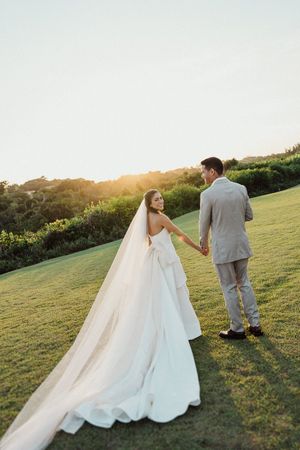 A couple in wedding attire walks on a grassy hillside at sunset with a long flowing veil trailing behind.