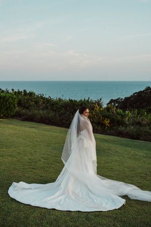 A bride in a flowing white wedding dress sits gracefully on lush green grass with the ocean visible in the background.