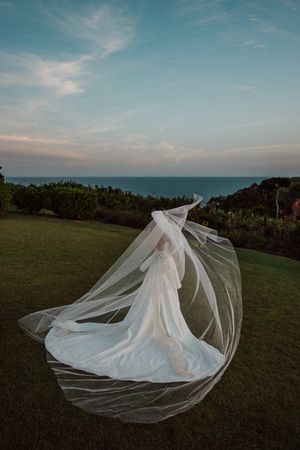 A flowing white wedding dress and veil billow dramatically on a hilltop overlooking the ocean at sunset.