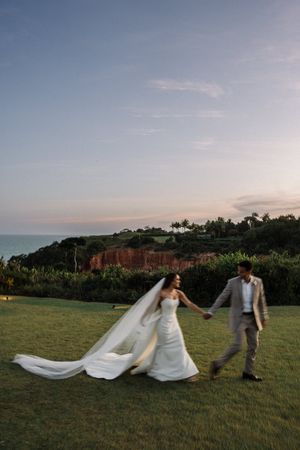Newlyweds walk hand in hand on a scenic cliff overlooking the ocean at sunset, with the bride's veil flowing in the breeze.