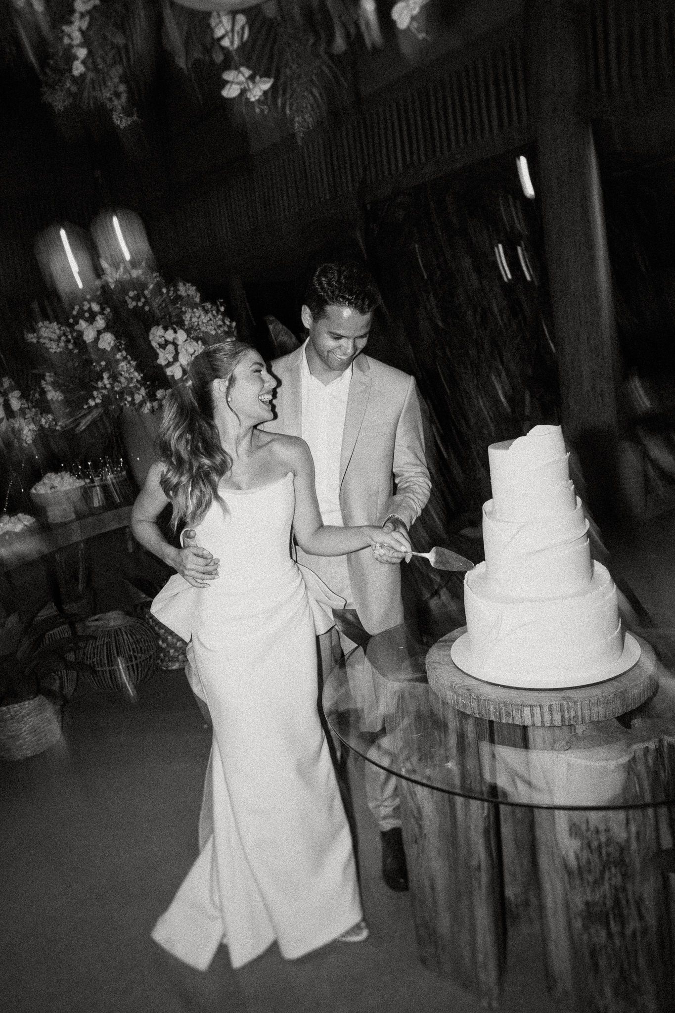 A bride and groom in black and white photography share a candid moment while cutting their elegant tiered wedding cake.