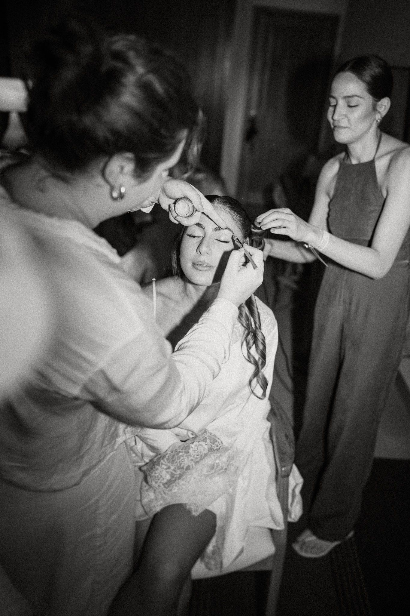 Black and white photo showing preparation before a wedding ceremony as helpers assist with styling and final touches.