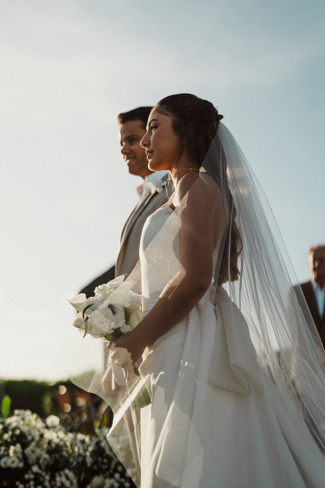 A bride in a flowing white dress and veil holds a bouquet while standing beside a groom during an outdoor wedding ceremony.