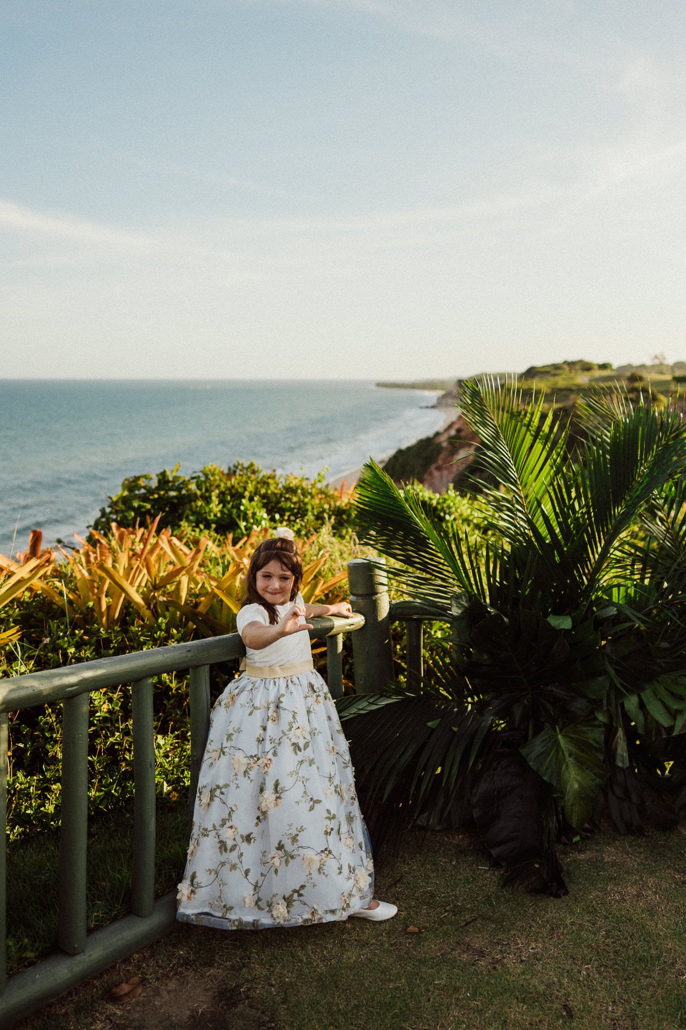 A child in a floral white dress stands on an oceanfront terrace with palm trees and tropical plants in Bali.