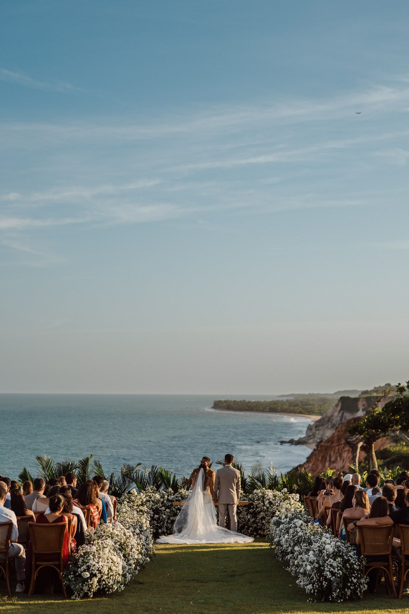 Wedding ceremony overlooking dramatic coastal cliffs at sunset with guests seated and white floral arrangements.