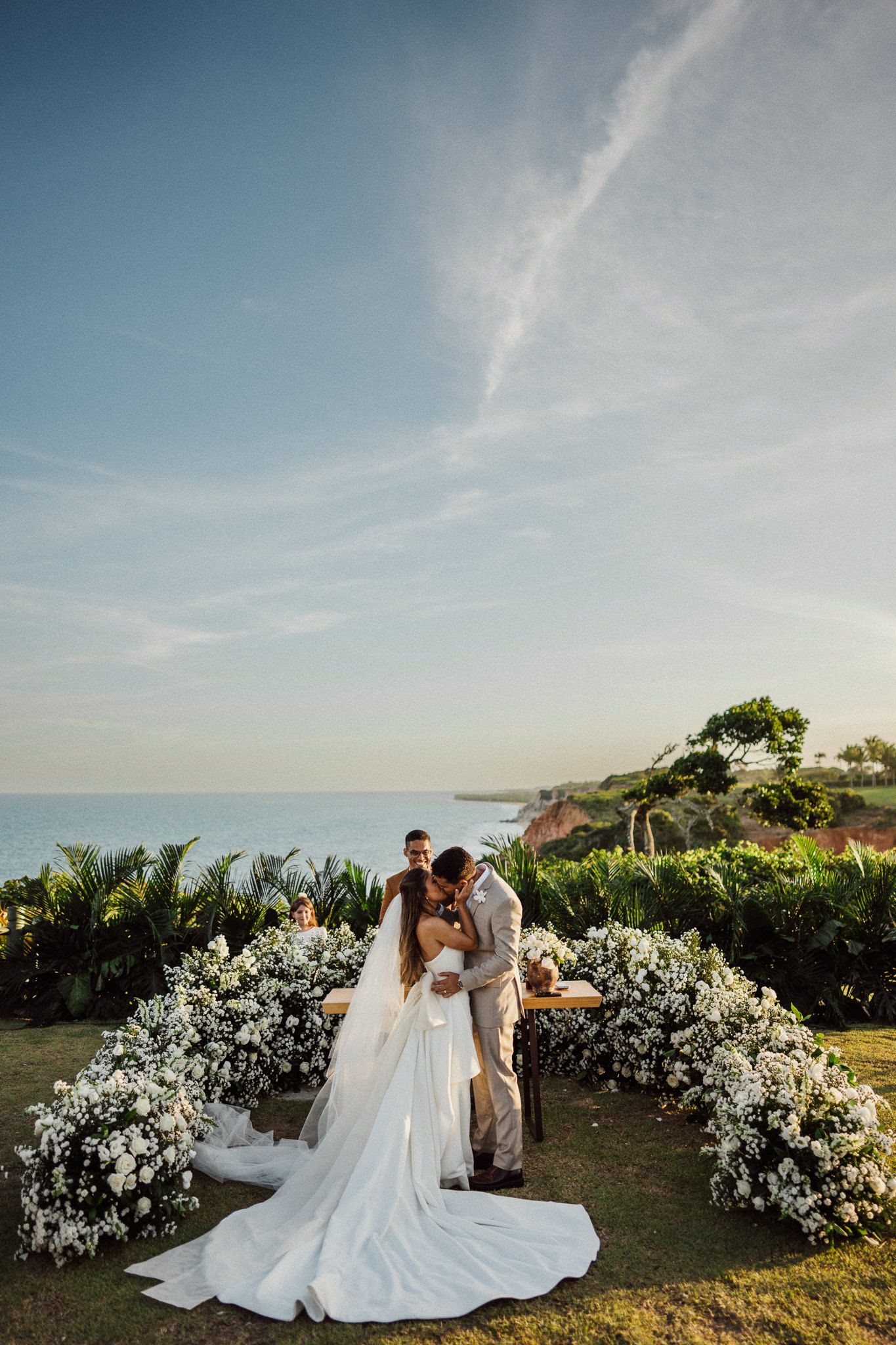 Romantic wedding photos on a coastal hillside with white floral arrangements at sunset.