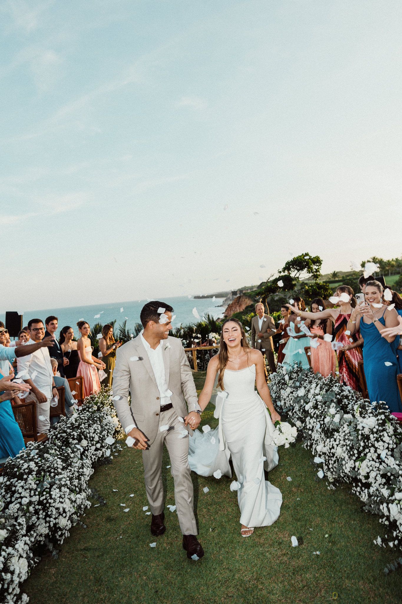 A joyful couple walks down an aisle lined with white flowers at their outdoor cliffside destination wedding ceremony.