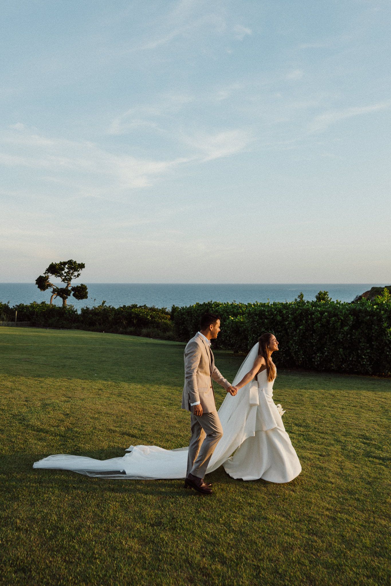 A couple in wedding attire walks together on a grassy hilltop with a beautiful sky during sunset.