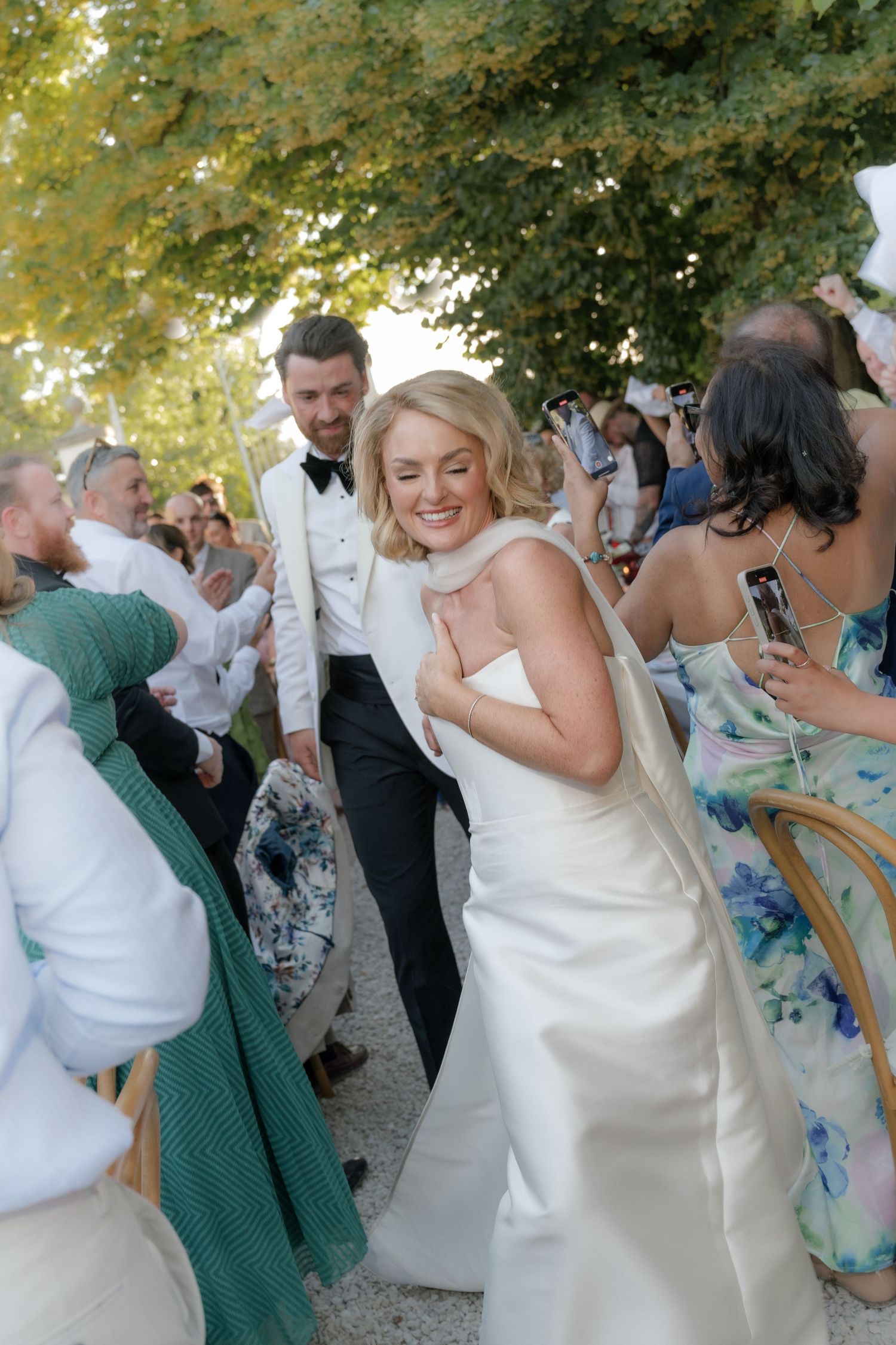 Celebratory wedding scene outdoors with white bridal gown and tuxedo amongst joyful guests under leafy trees.