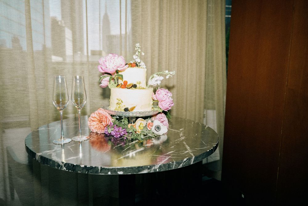 White wedding cake decorated with pink and purple flowers on a mirrored table with champagne flutes.