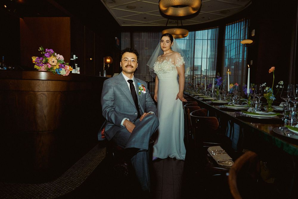 A wedding couple poses by an elegantly set dinner table in a dimly lit venue with floral centerpieces.