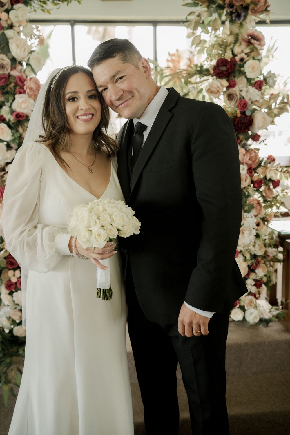 A wedding couple poses in front of a floral arch with white and burgundy flowers.