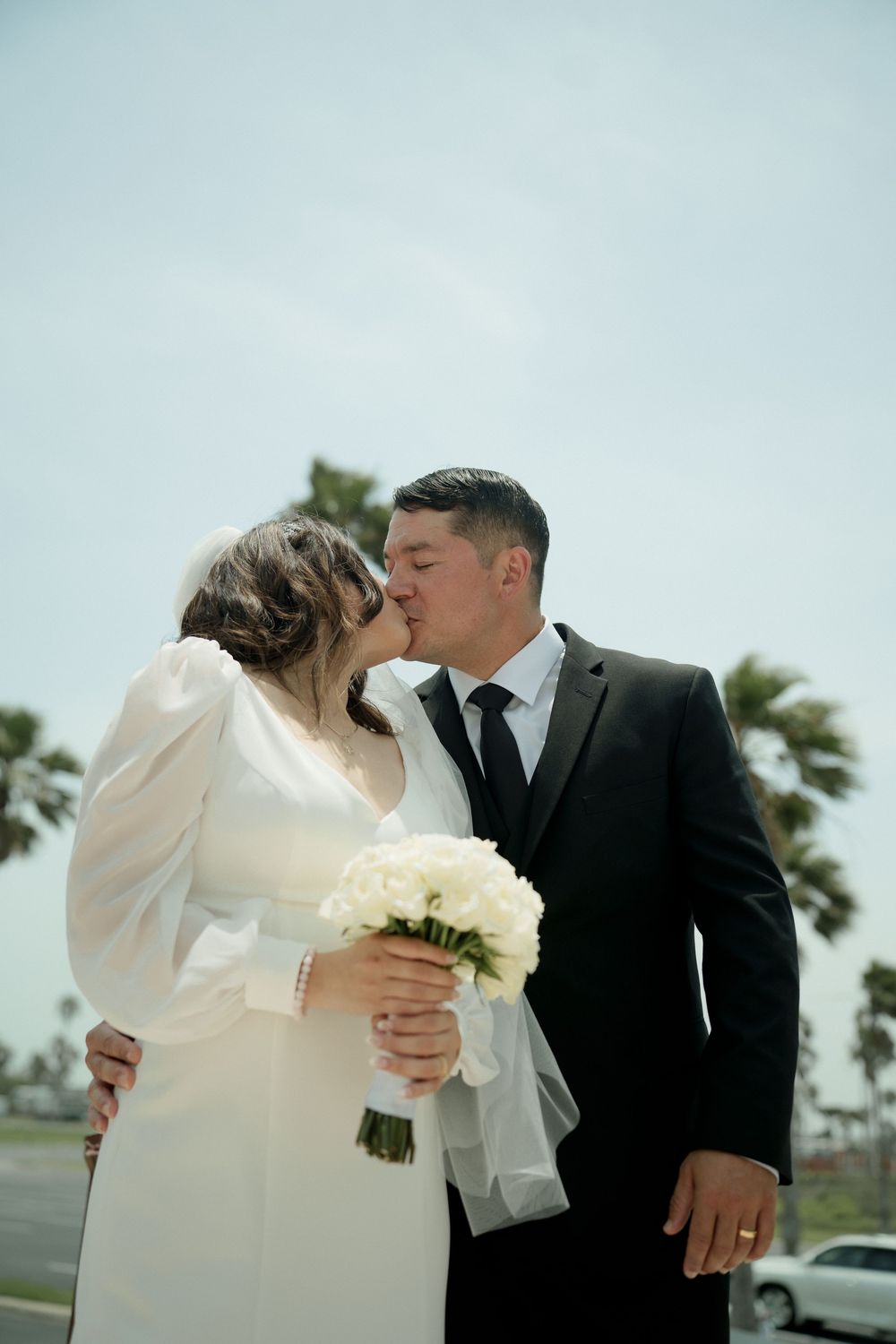 A romantic wedding kiss captured outdoors with palm trees in the background.