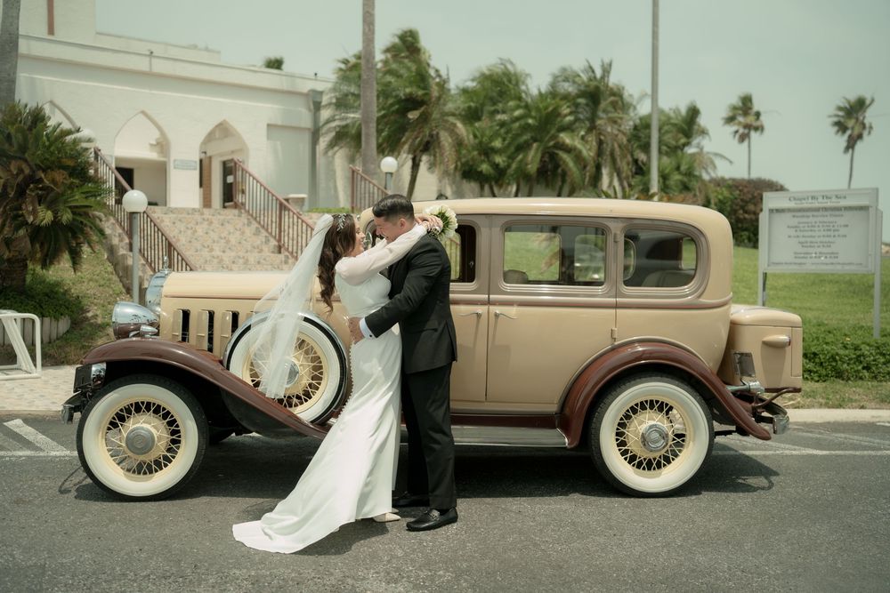 Newlyweds pose beside a vintage beige car outside a white church building.