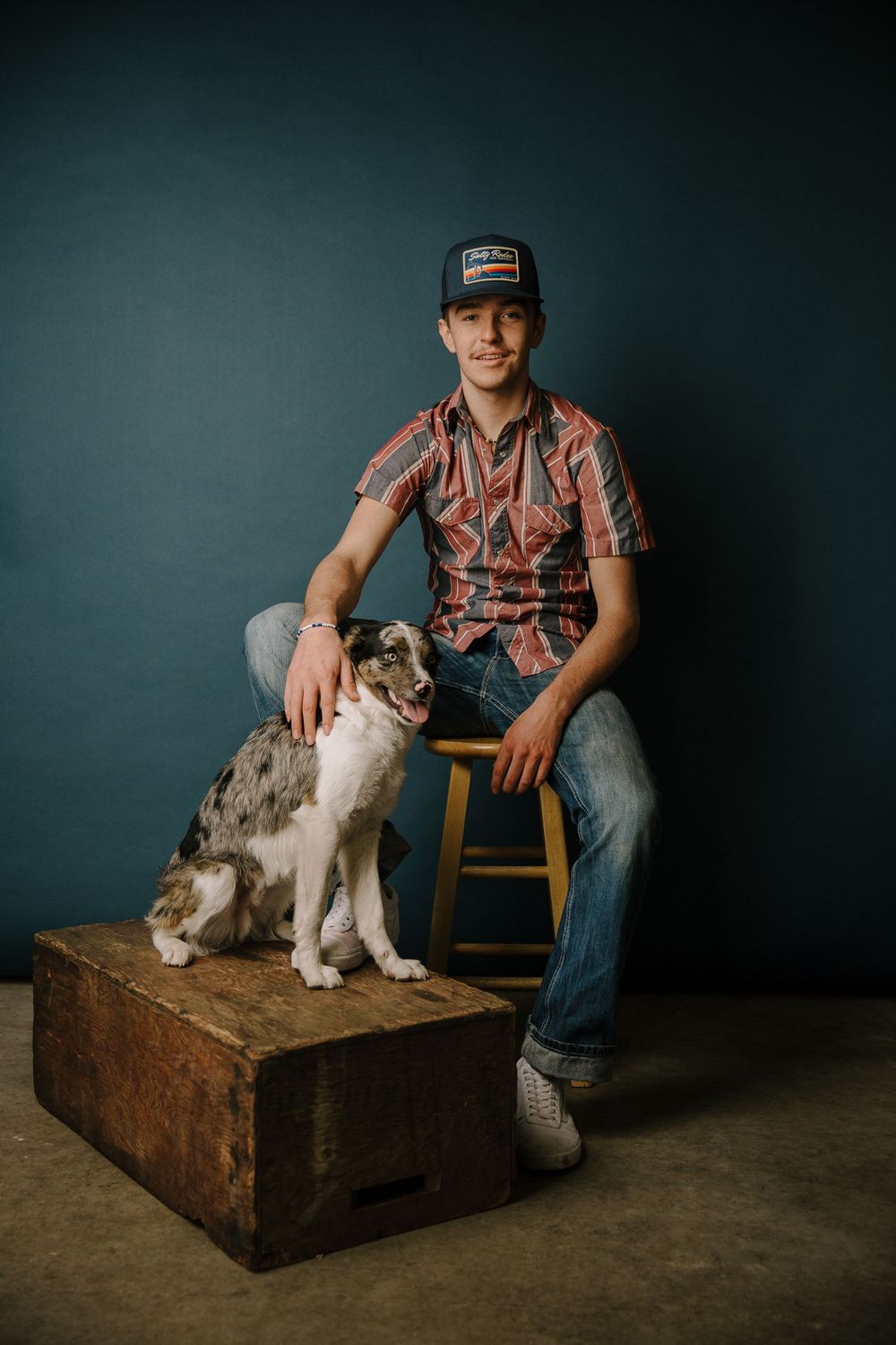 Person in casual attire sits with a spotted dog on a wooden platform against dark backdrop.