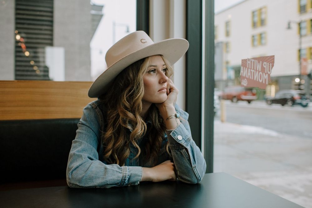 Person wearing hat gazes thoughtfully through window in moody cafe setting.