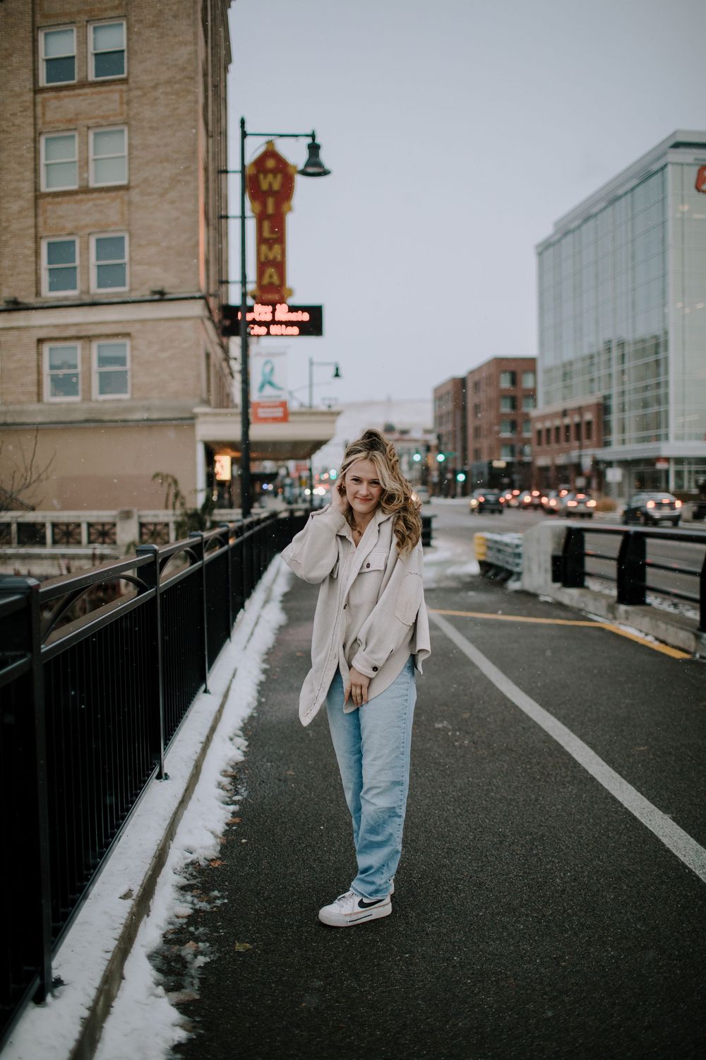 Person in beige coat and jeans walking on an urban street with buildings in the background.