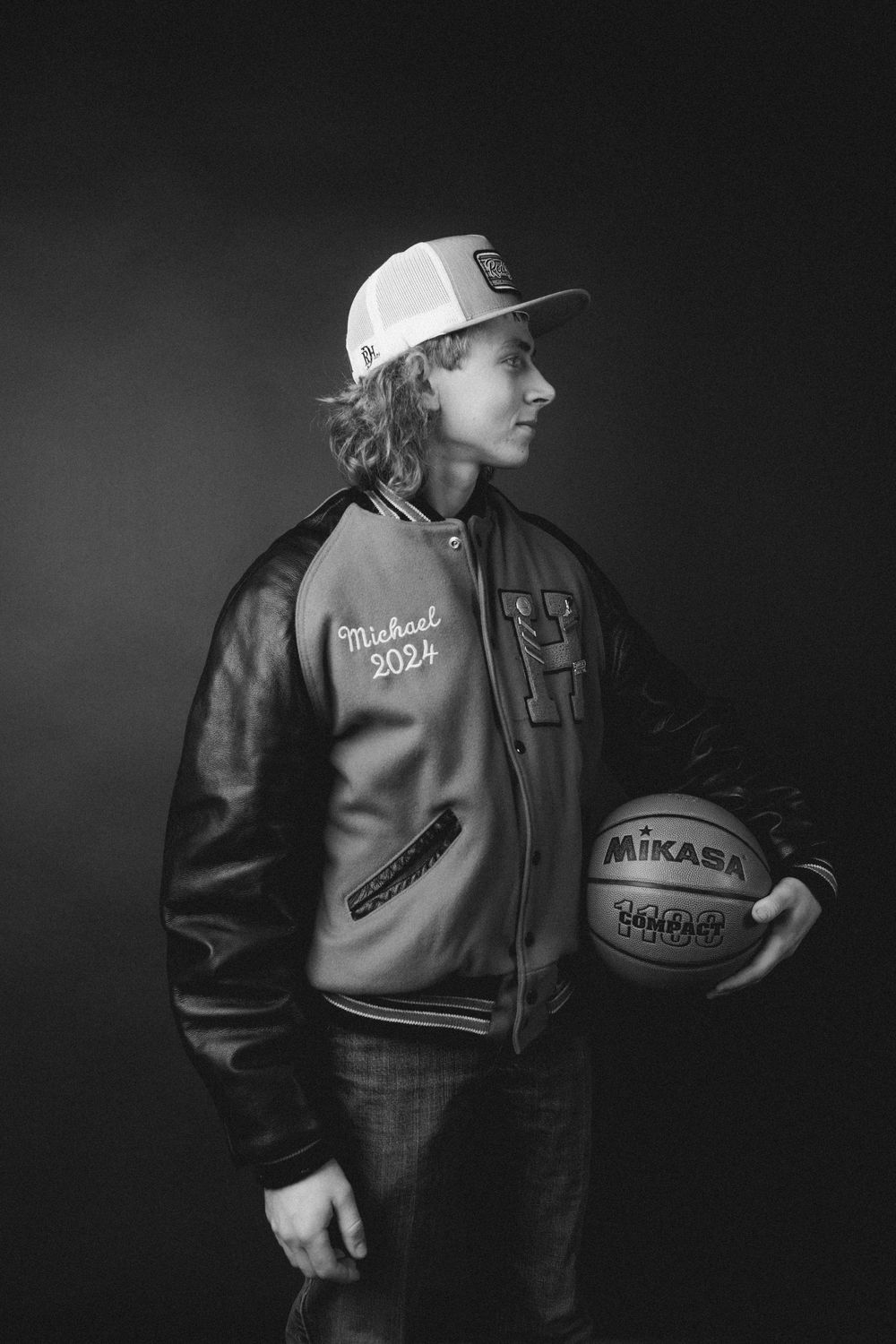 Black and white portrait with a basketball and varsity jacket against dark background.
