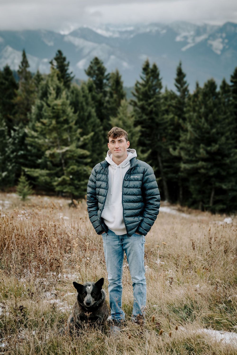 Person in puffy blue jacket and jeans poses in mountain field with evergreen trees and snowy peaks in background.